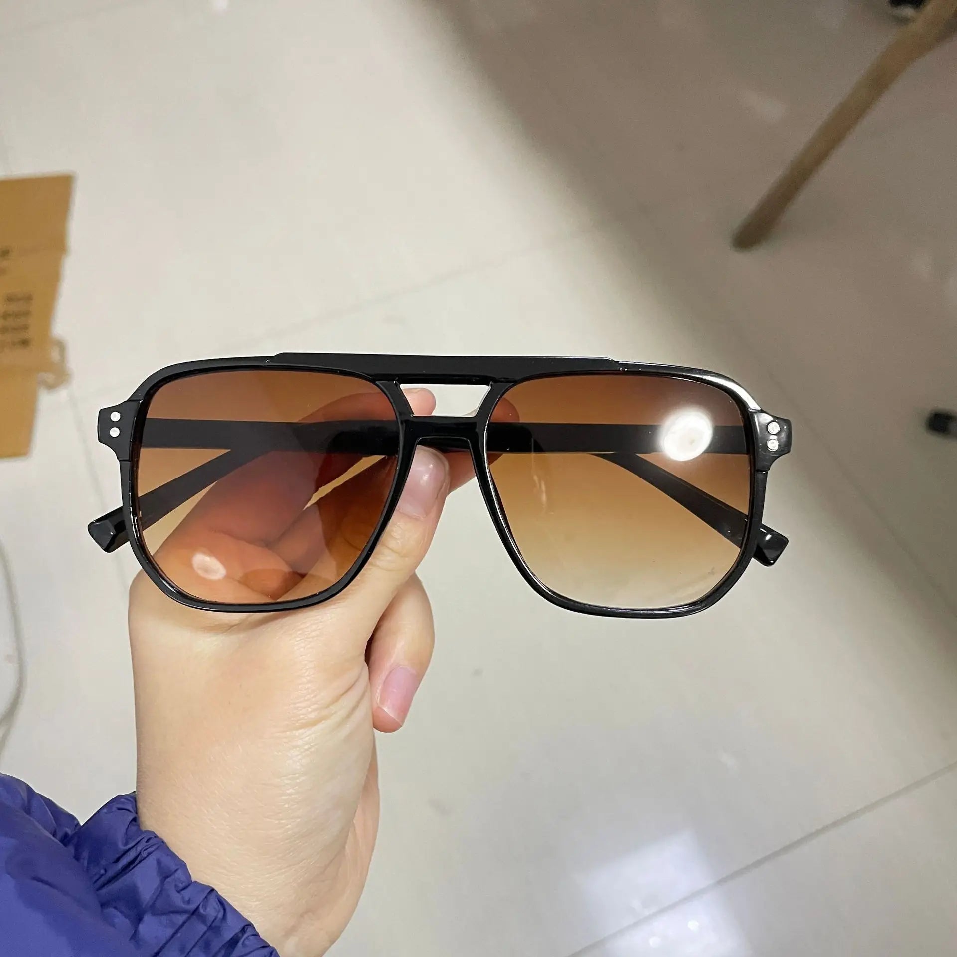Sunglasses with black frame and brown lenses held by a hand against a neutral background